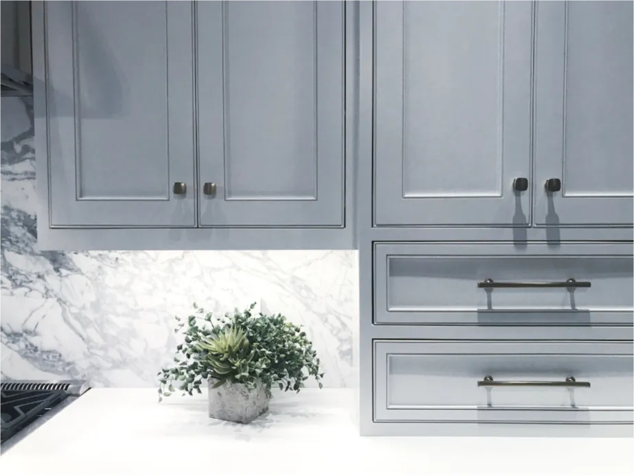 Cabinet painting - Gray kitchen cabinets with brass handles, marble backsplash, and a potted plant on the white countertop.