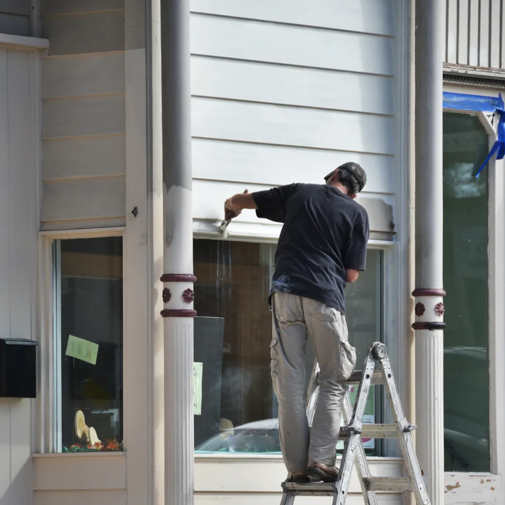 commercial-painting Person on ladder painting white window frame of a building with two large windows.