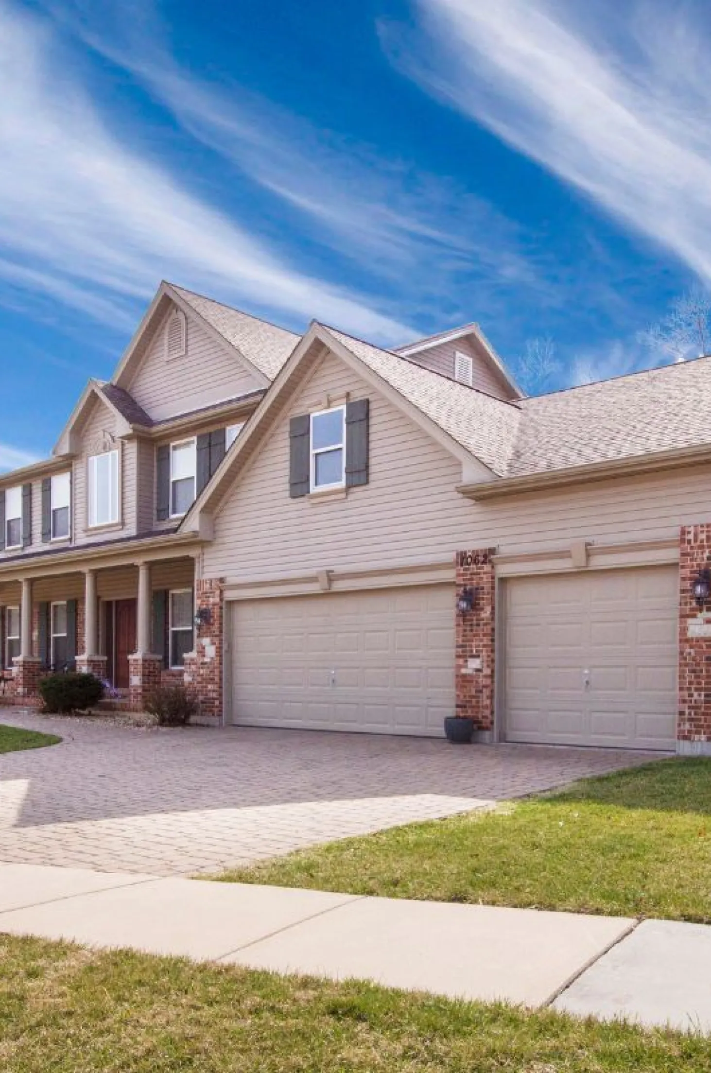 exterior-painting-3 Two-story suburban house with a three-car garage, brick accents, and a cloudy blue sky.