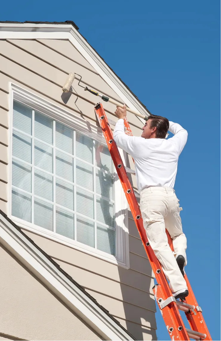 exterior-painting-4 Person on a ladder painting the exterior of a house near a window against a clear blue sky.