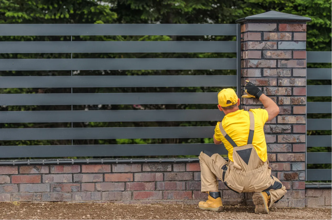 fence-building-1 A worker in yellow and brown overalls installs a metal fence on brick columns outdoors.