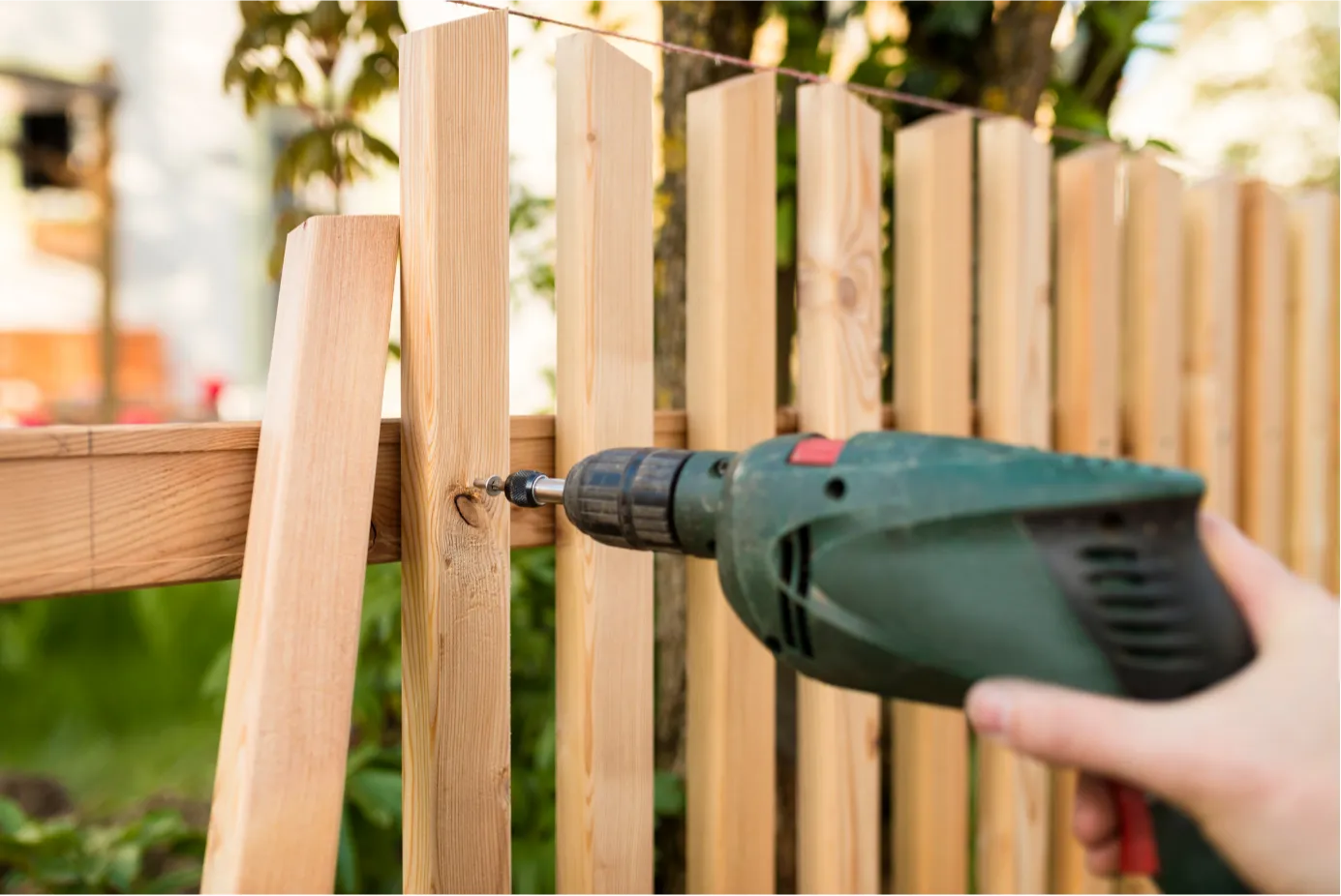 Close-up of a person using a green power drill to fasten wooden planks on an outdoor fence.