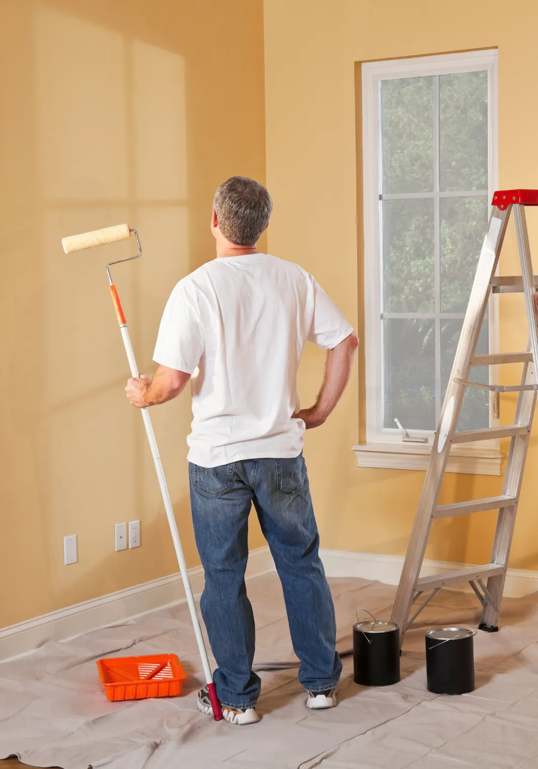 interior-painting-3 Person holding a paint roller, standing in a room with yellow walls, next to a ladder and paint cans, facing a window.