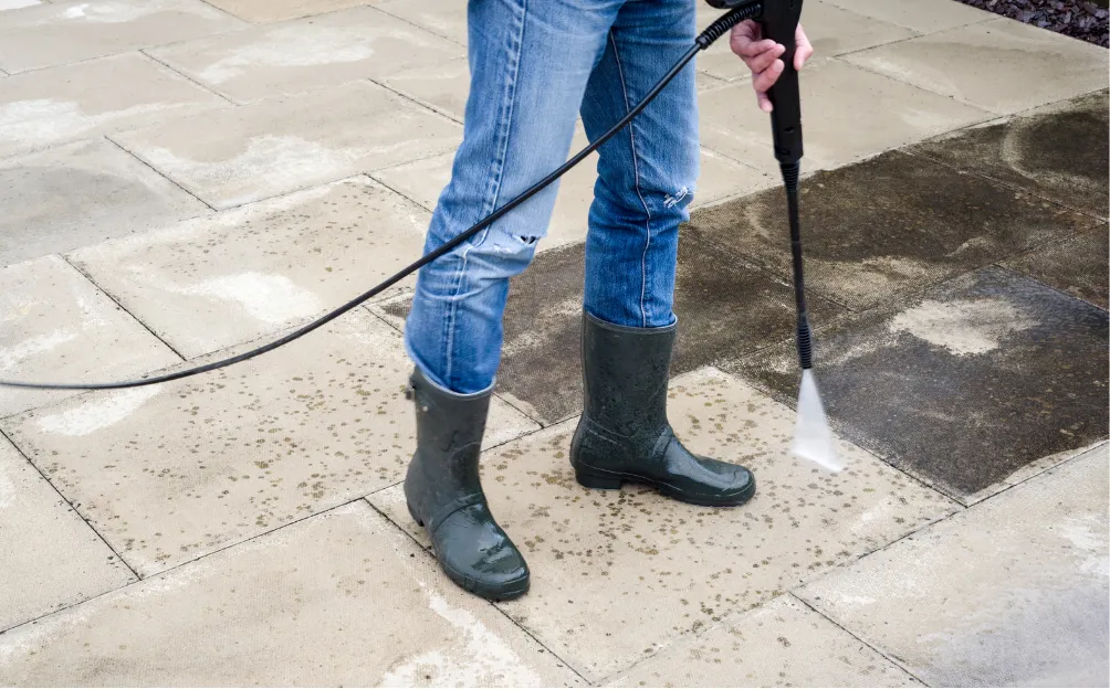pressure-washing-4 Person in jeans and boots power washing a dirty patio, revealing clean tiles beneath.