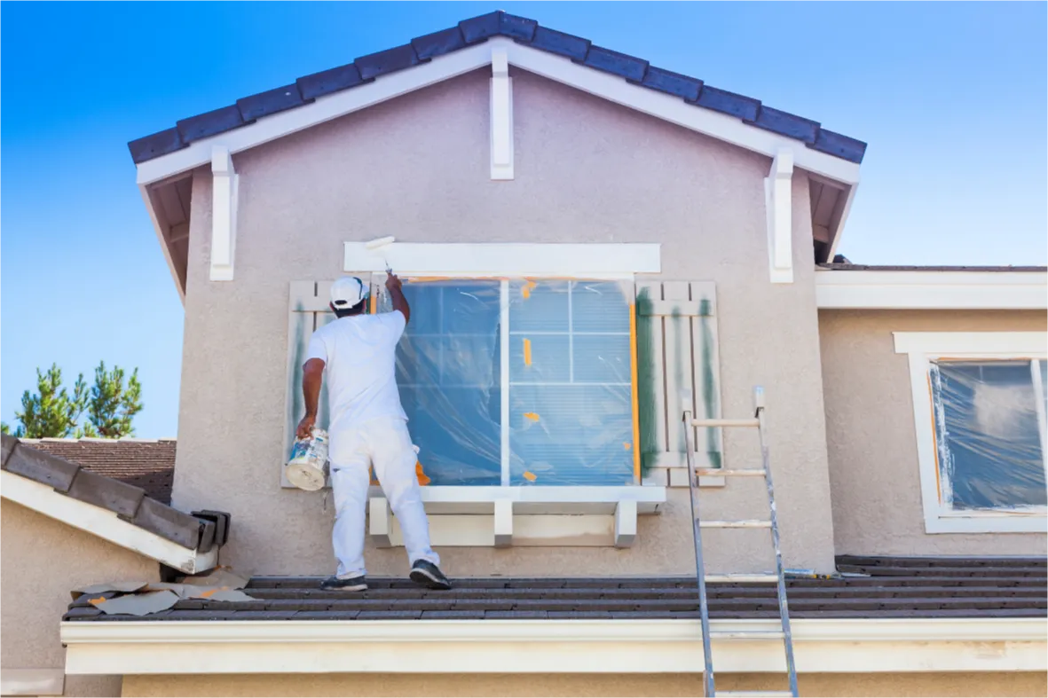 residential-painting-1 A painter in white overalls paints the exterior of a house while standing on the roof beside a ladder.