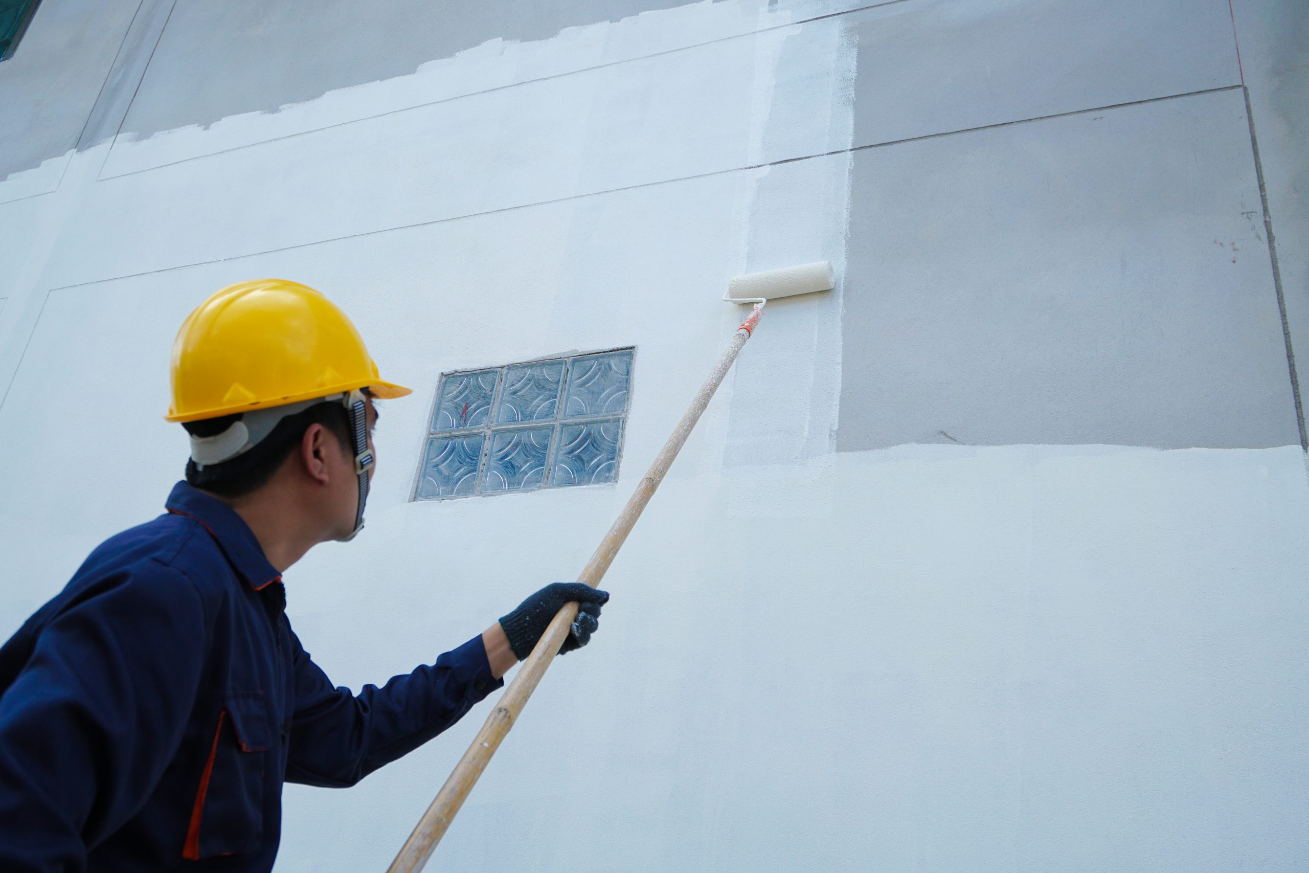 Asian male construction worker is painting with paint roller on white house wall, home decoration and construction concept in project. A worker in a yellow hard hat paints a wall white with a roller on a long pole.